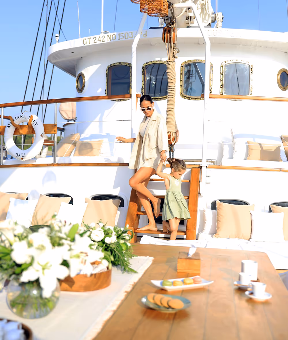Mother and child stepping down a wooden staircase on a luxury yacht in bright sunlight, with a table set for afternoon tea in the foreground and white cushions and floral arrangements decorating the deck.