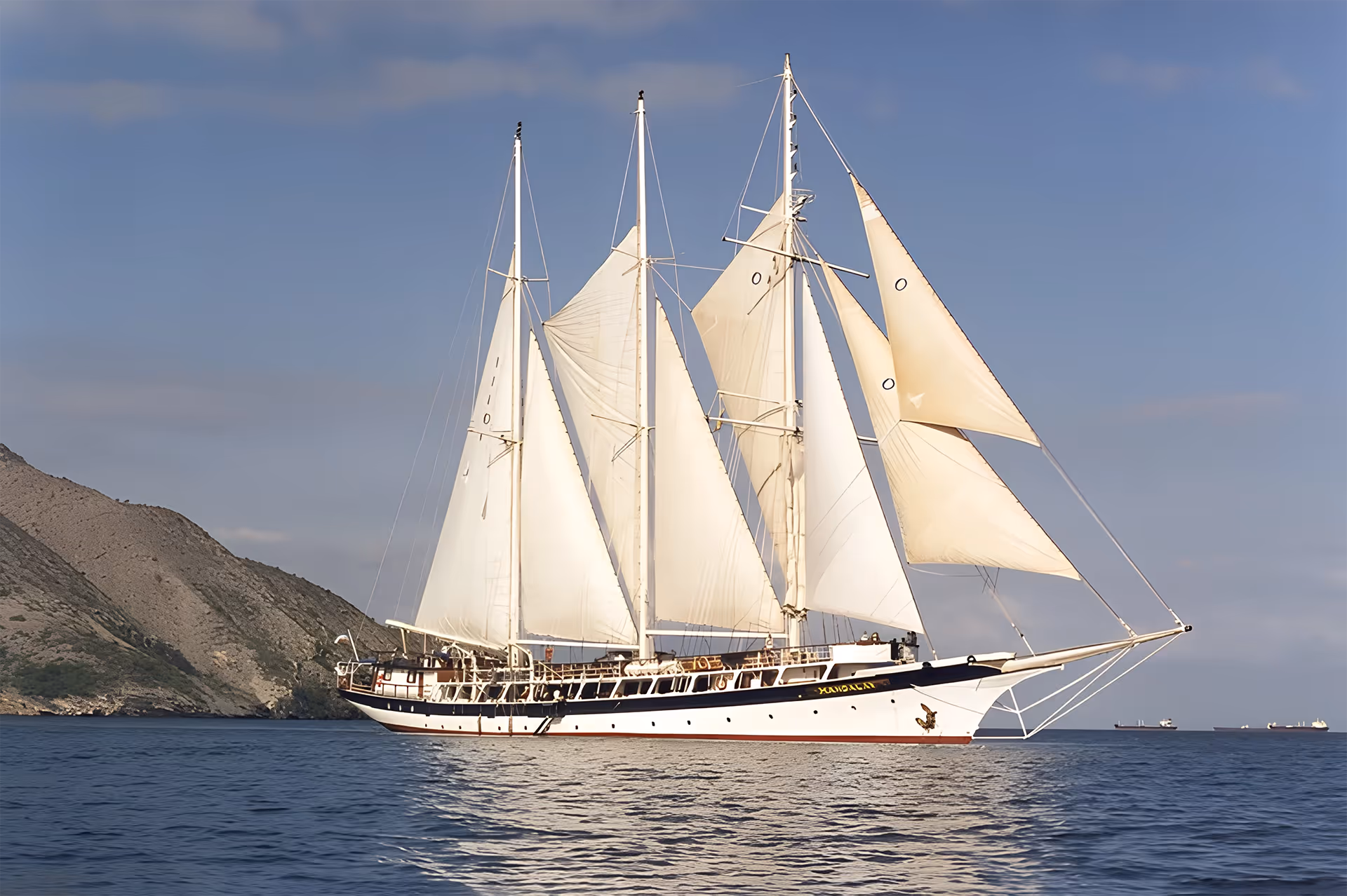 Three-masted sailing schooner with full white sails gliding across calm blue waters near a rocky mountainous coastline under a clear sky.
