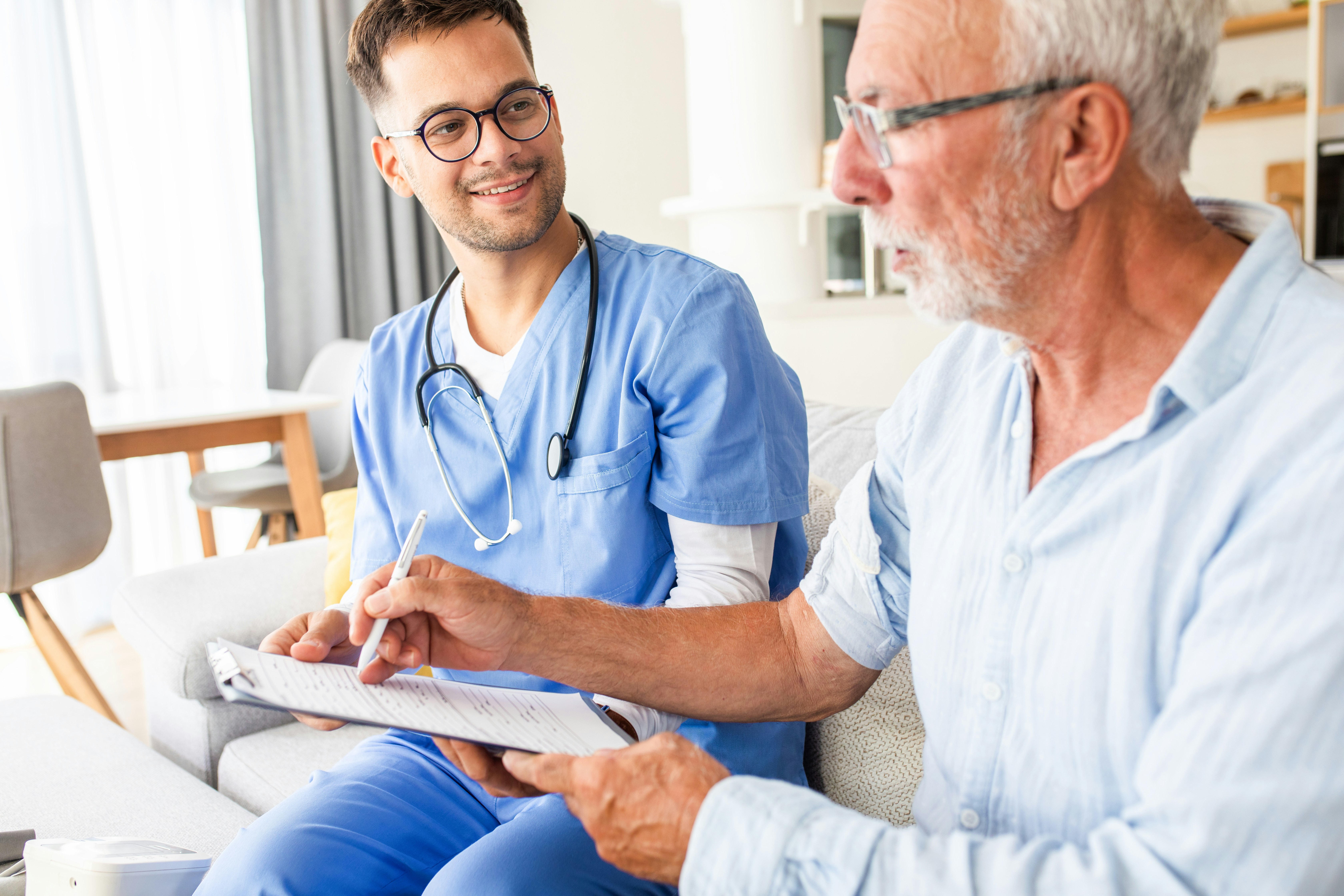 Young male nurse in blue scrubs and glasses consulting with elderly man holding a pen and form in a living room.