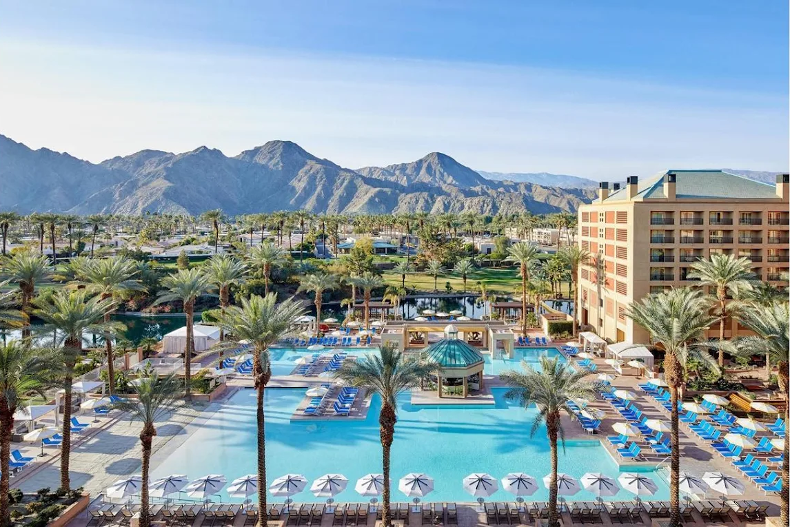 Large resort swimming pool surrounded by palm trees, blue lounge chairs, white umbrellas, and a multi-story hotel building with mountains in the background under a clear blue sky.