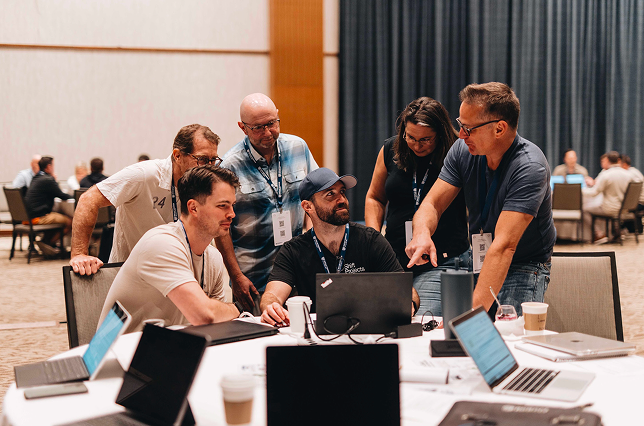 Group of five men wearing conference badges gathered around a laptop, smiling and looking engaged.
