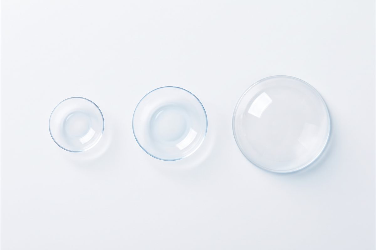 Three contact lenses side by side on a white surface showing their different sizes: a small rigid gas permeable lens, a soft lens and a large scleral lens.