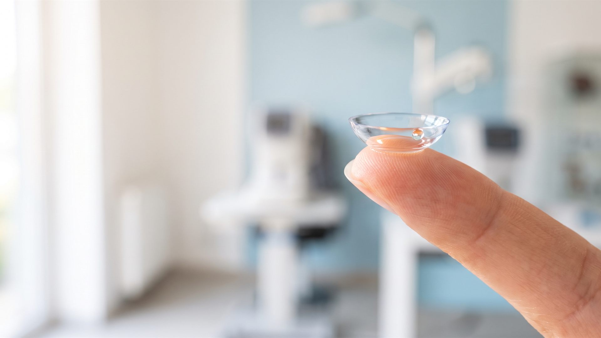 A clear scleral contact lens balanced on a fingertip in a bright optometry clinic.