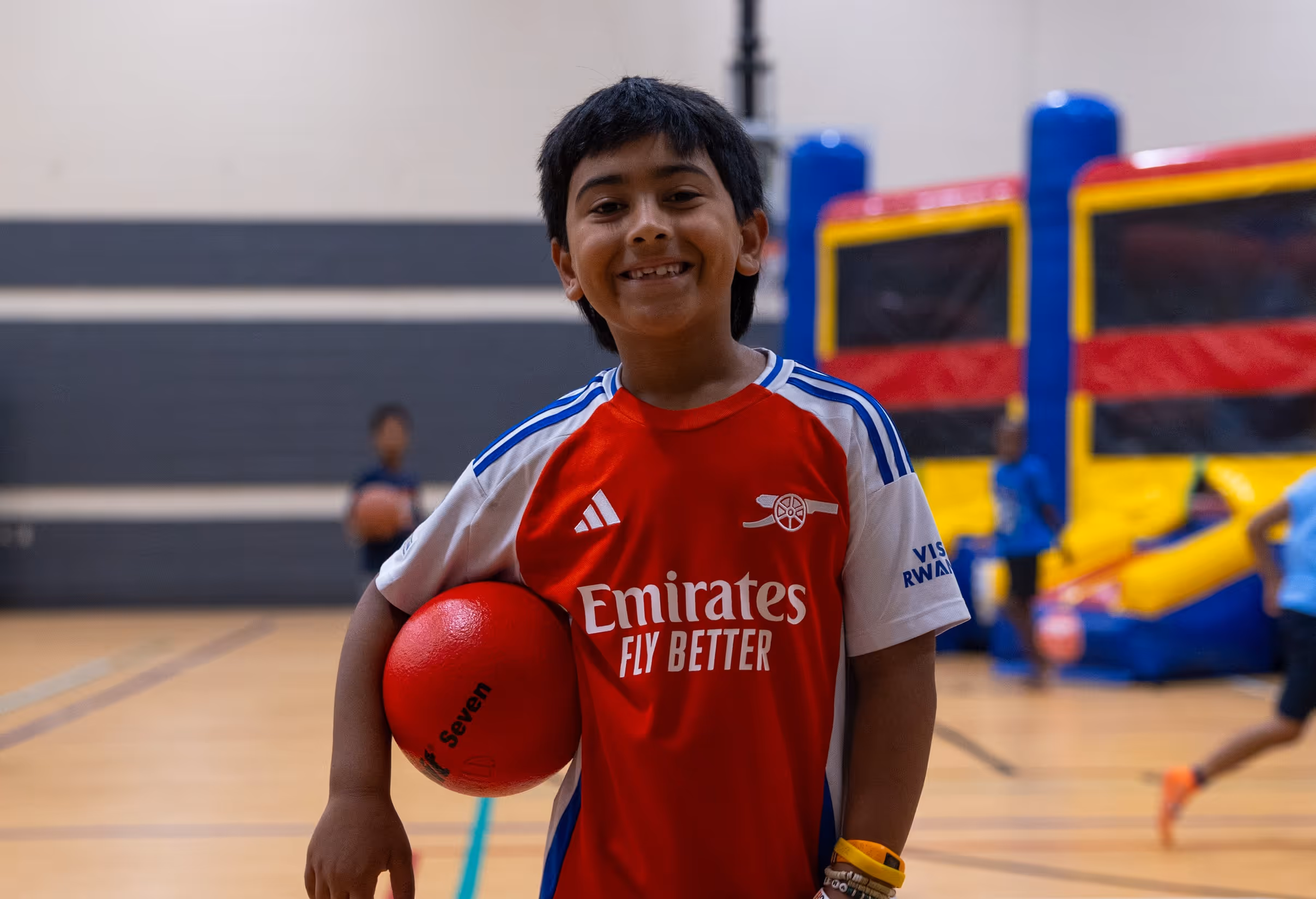 Smiling boy in a red and white Arsenal football jersey holding a red ball at summer day camp with children and inflatable play equipment in the background.