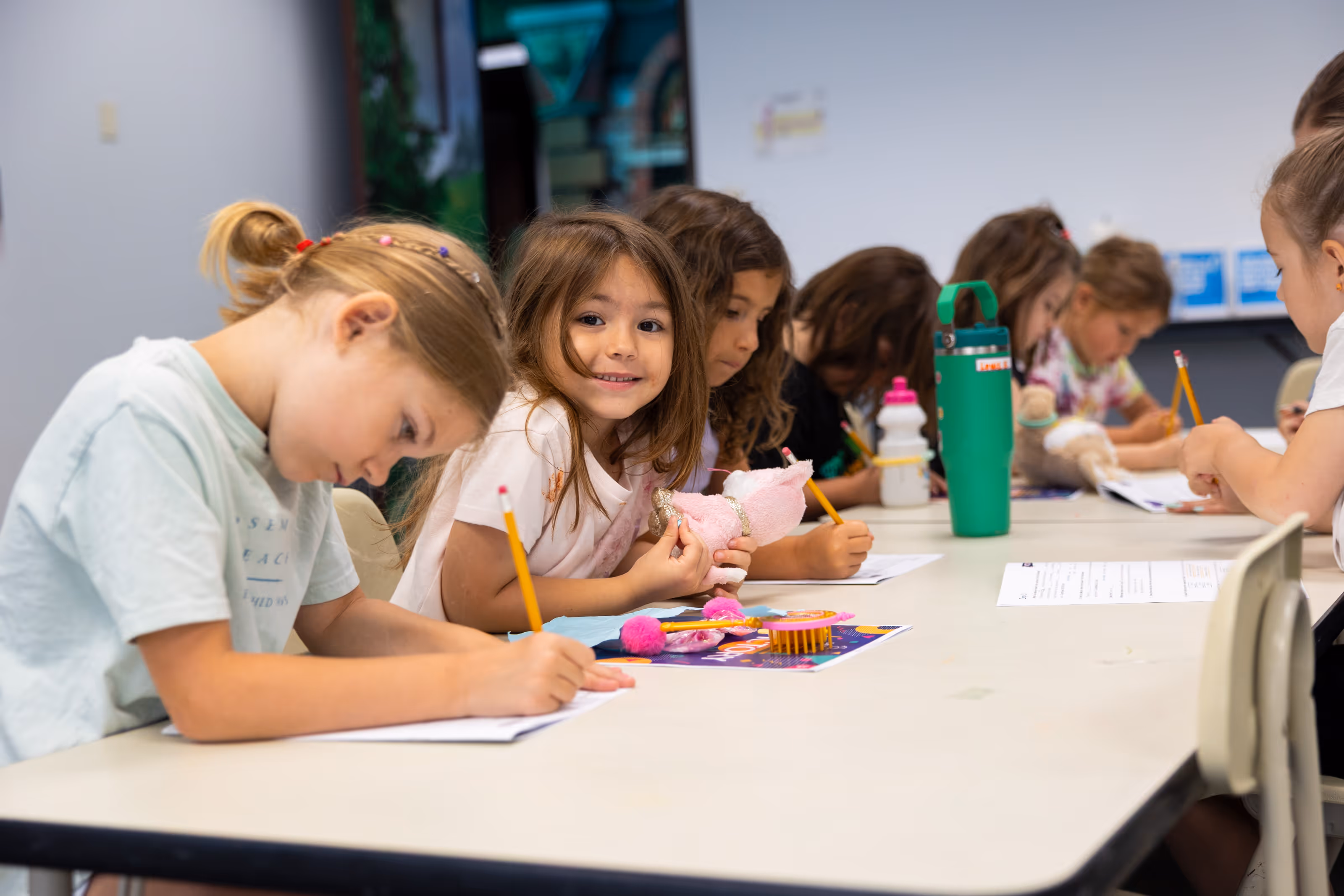 Group of young children sitting at a table at summer camp, writing with pencils on paper, one girl smiling while holding a pink stuffed animal.