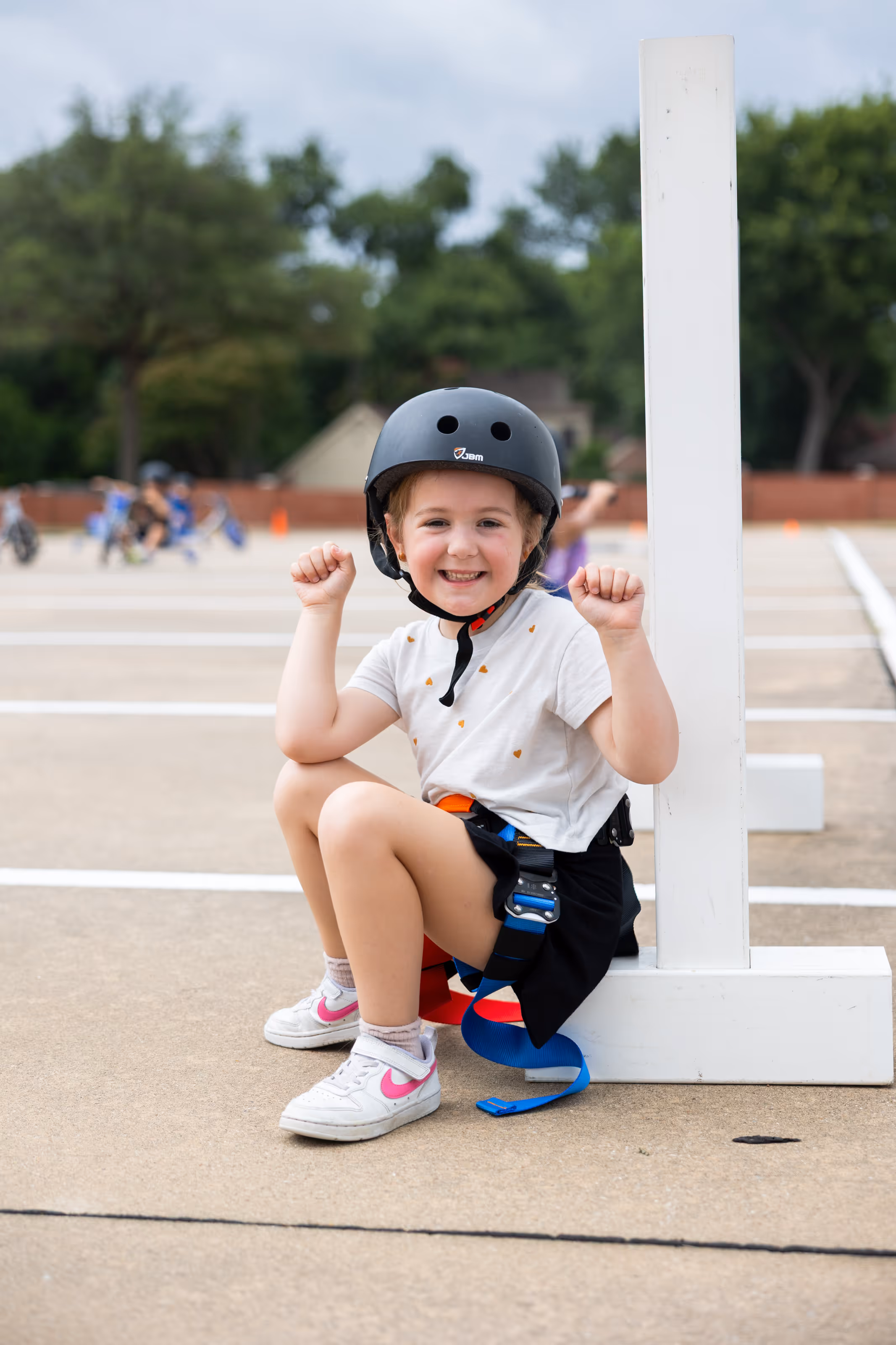 A smiling young girl wearing a helmet and safety harness crouches next to a white post in an outdoor parking lot at Skylark summer day camp.