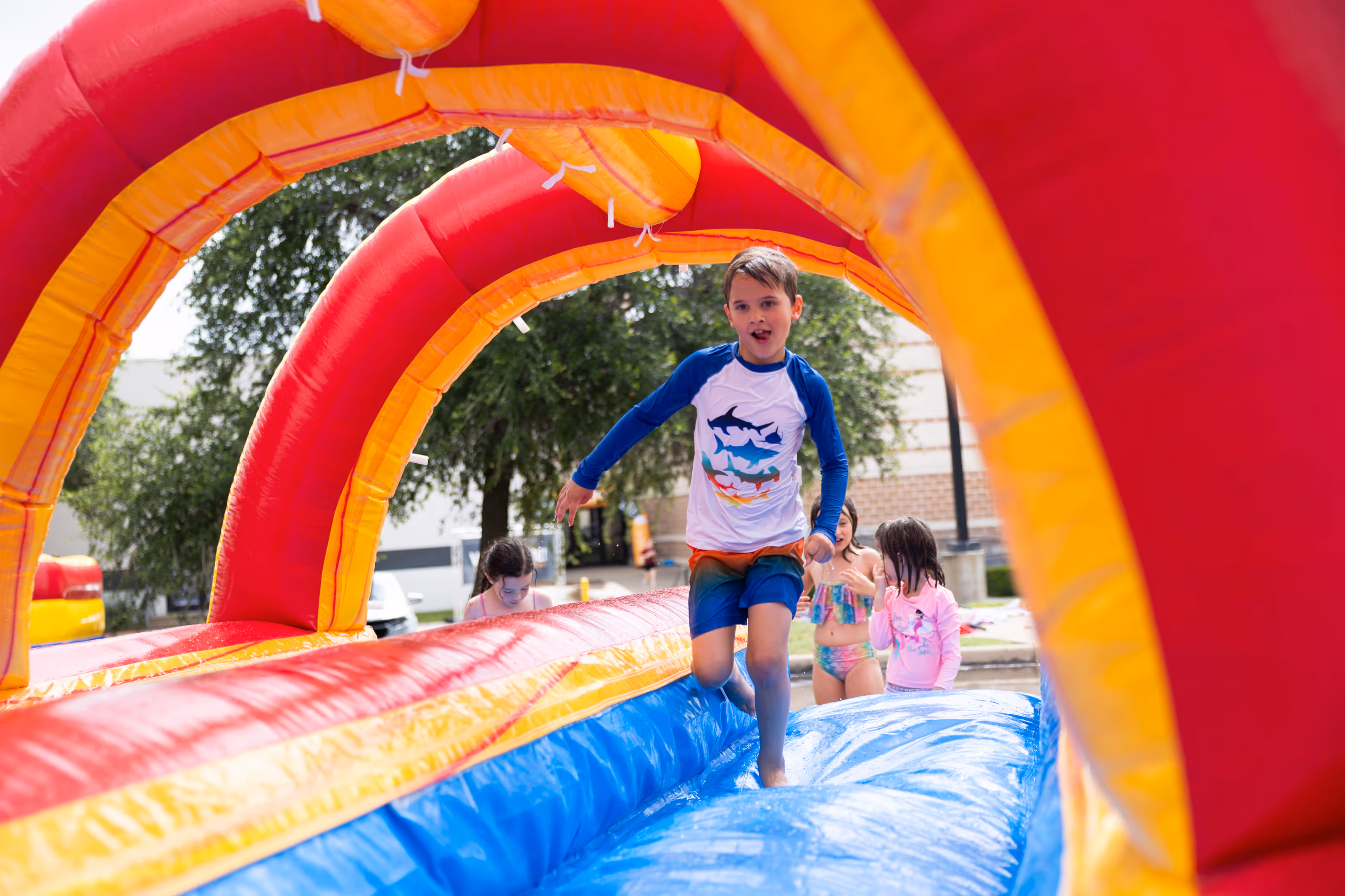 Boy in a white and blue shark-themed rash guard running through a colorful inflatable water slide tunnel with three girls in swimsuits standing behind him at summer camp.