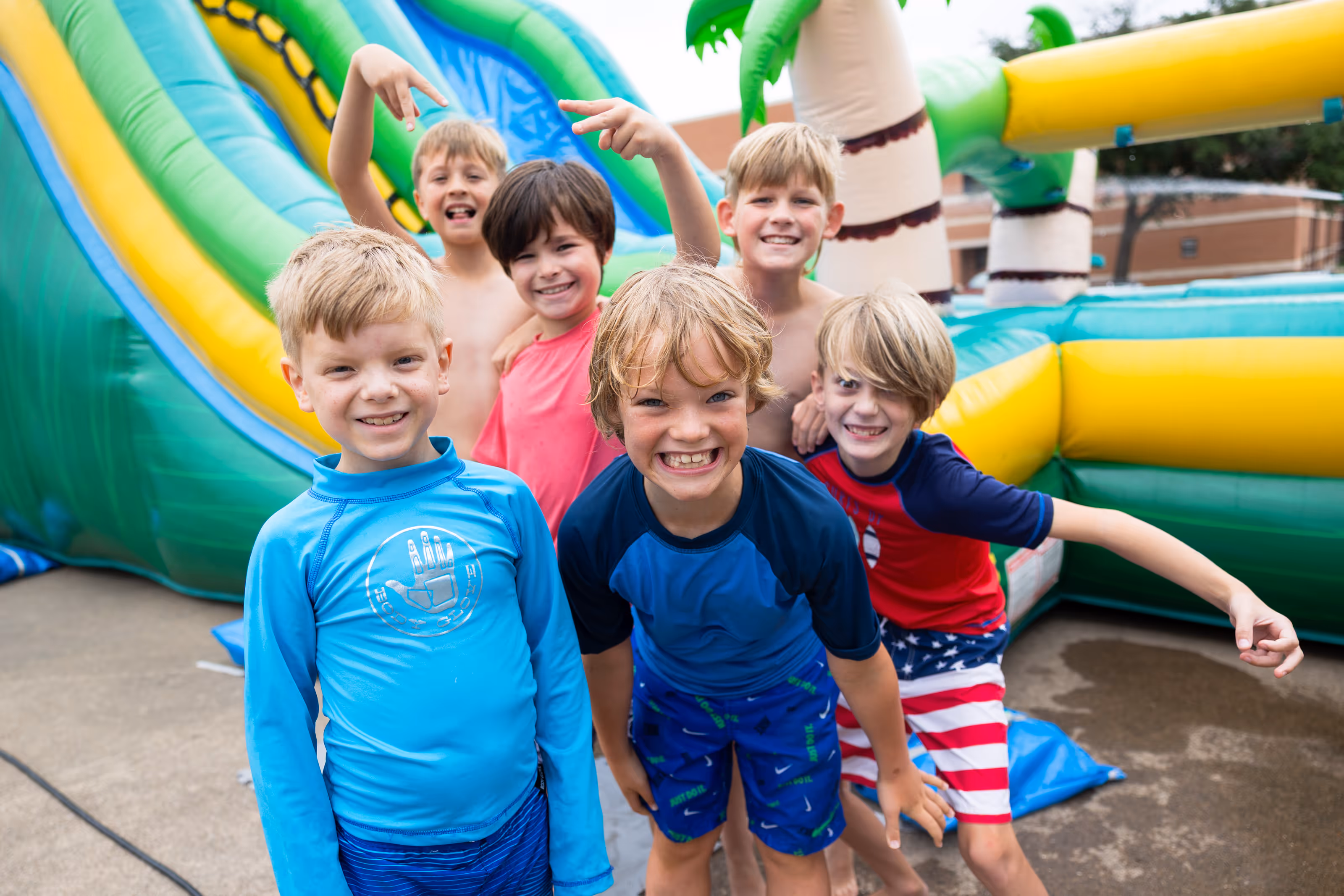 Six smiling boys in swimwear posing happily in front of a colorful inflatable water slide at summer camp.