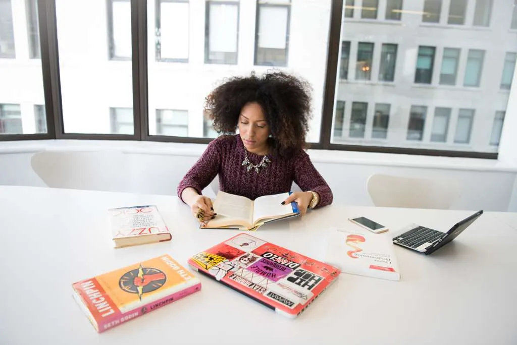 Woman with curly hair reading a book at a white table with books, a laptop, and a smartphone.