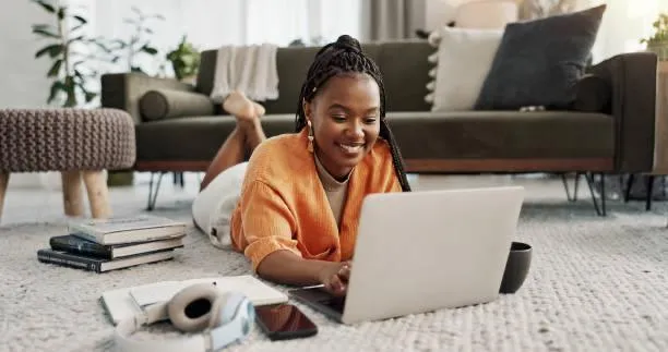 Smiling woman lying on carpet in living room using a laptop with books, headphones, and smartphone nearby.