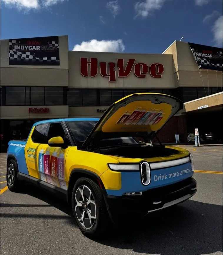 Yellow and blue branded electric pickup with its front trunk open outside a Hy-Vee store