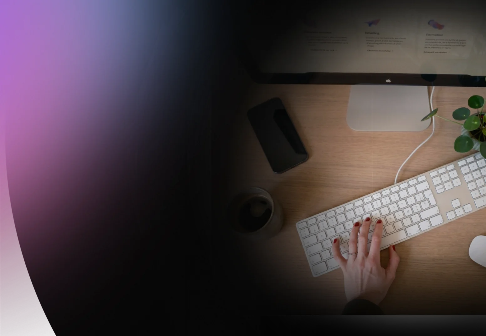 Main droite tapant sur un clavier blanc d’ordinateur sur un bureau en bois avec un écran, un téléphone portable, une tasse et une plante verte.