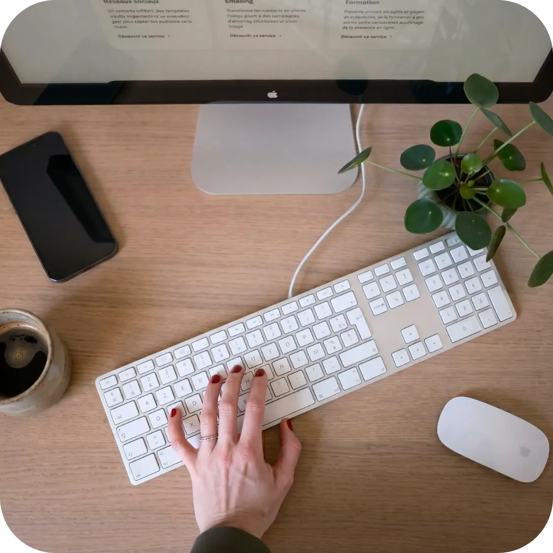 Une main avec des ongles vernis de rouge tapant sur un clavier blanc d'ordinateur posé sur un bureau en bois, avec un écran, un téléphone, une tasse de café, une souris et une plante verte.