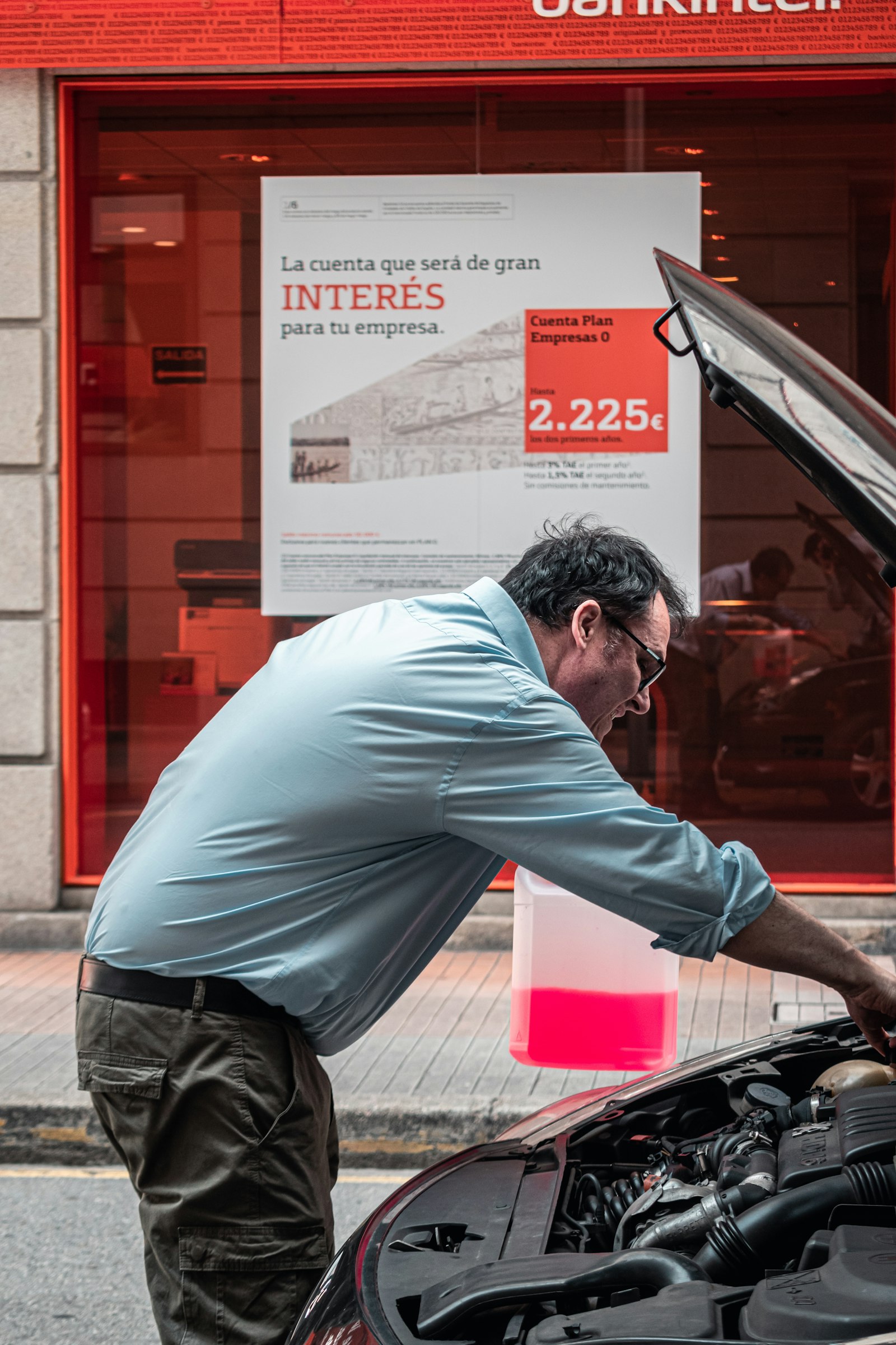 Man inspecting a car engine on a Spanish city street, Bankinter bank advertisement visible in the background