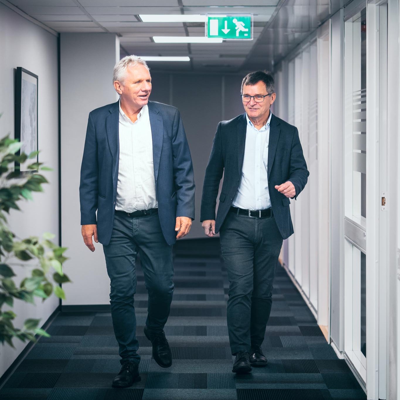 To menn i dress går nedover en kontorkorridor og smiler mens de snakker sammen.