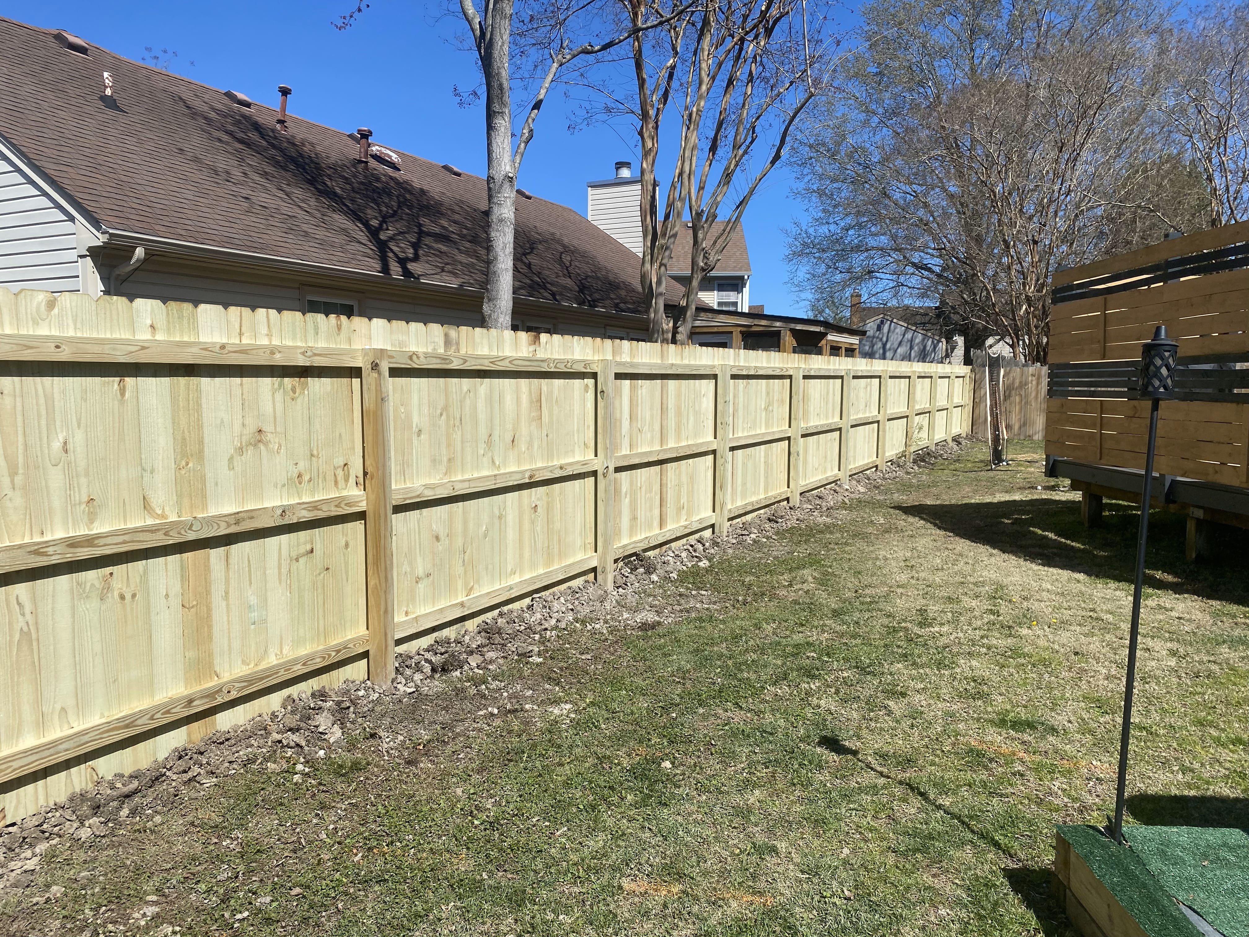 Long new wooden privacy fence running alongside a grassy backyard with houses and trees in the background under a clear blue sky.