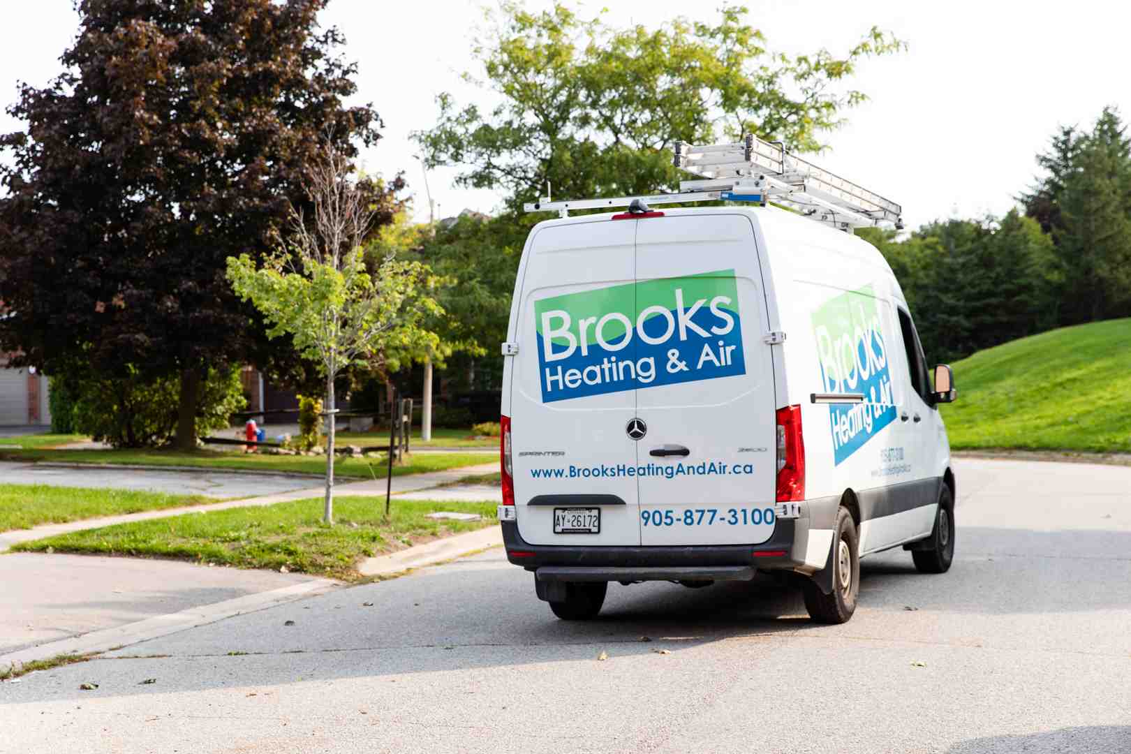 A professional white service van with a ladder rack parked on a snowy residential street in Acton ON during a bright winter day, ready for an emergency heating call - heating repair in acton on