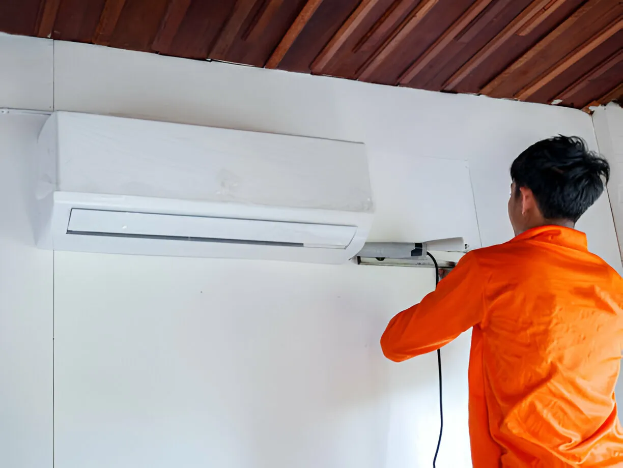 Technician in orange shirt installing or repairing a white wall-mounted air conditioner