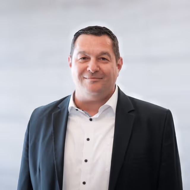 Mann mittleren Alters mit kurzen dunklen Haaren, in weißem Hemd und dunklem Anzug vor unscharf hellem Hintergrund.