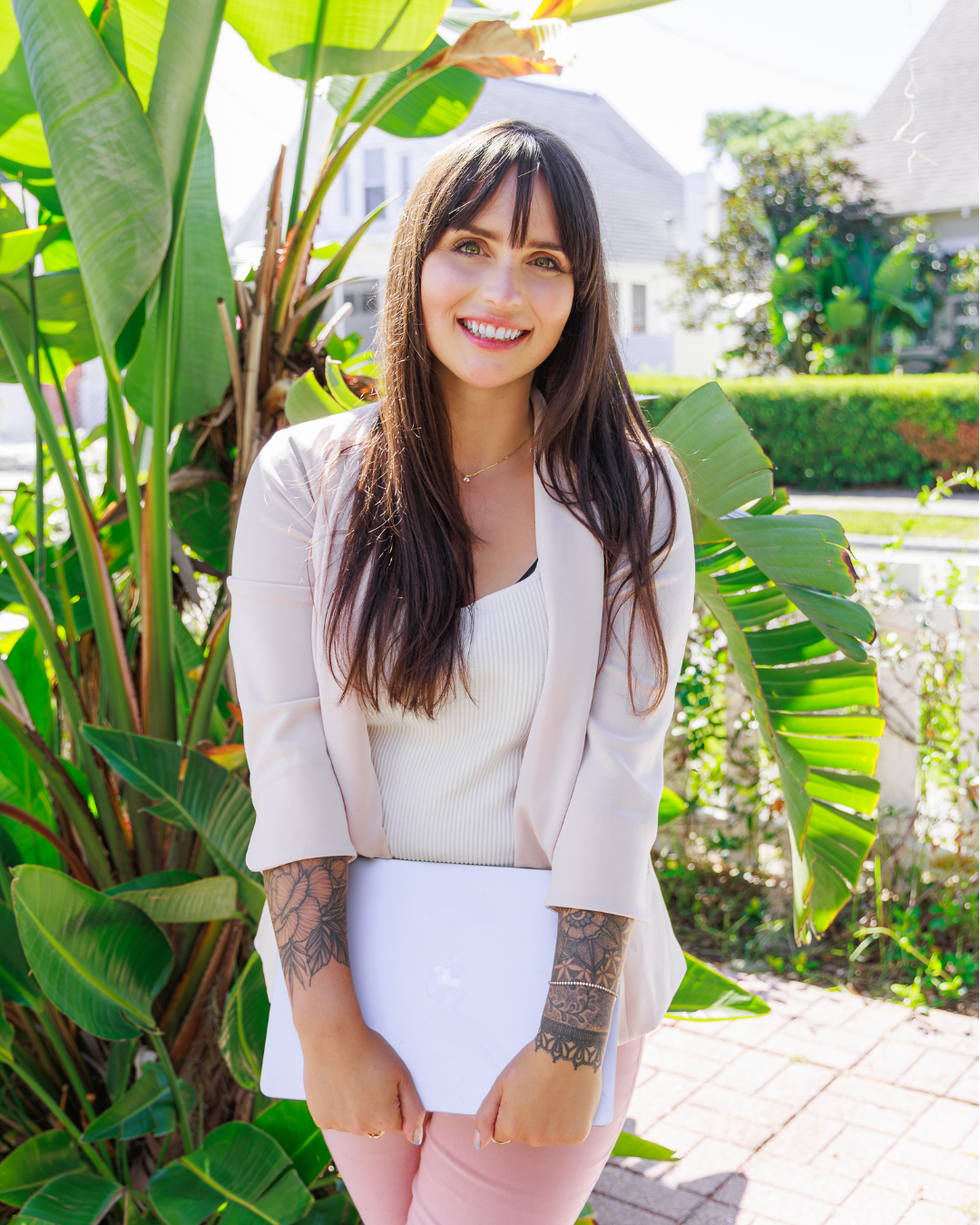Professional brand portrait of Amanda Sinnott (Hello Edu) smiling, standing outdoors with bright tropical foliage (like a Bird of Paradise plant) behind her, holding her laptop.