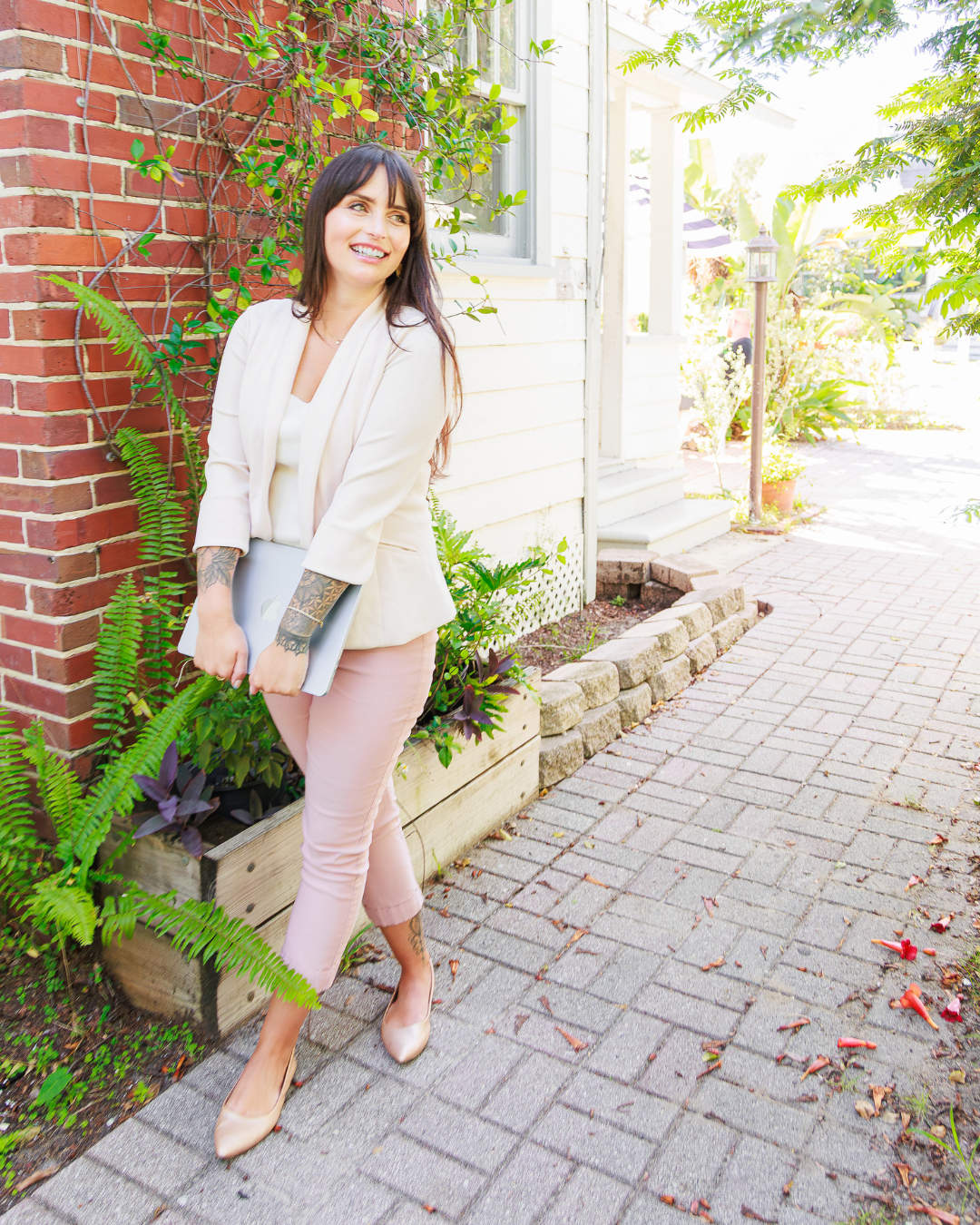 Full-body brand portrait of Amanda Sinnott, Hello Edu, standing outdoors next to a brick wall covered in ivy. She is wearing a blazer and pink pants, holding her laptop, and looking confidently into the distance.
