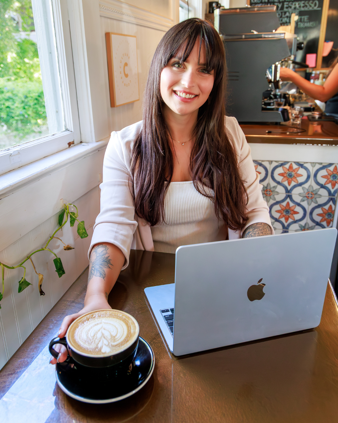 Amanda Sinnott, founder of Hello Edu, smiling and working on her MacBook at a cafe table, holding a cup of latte art coffee. Professional brand portrait showing an educational consultant in a work environment.