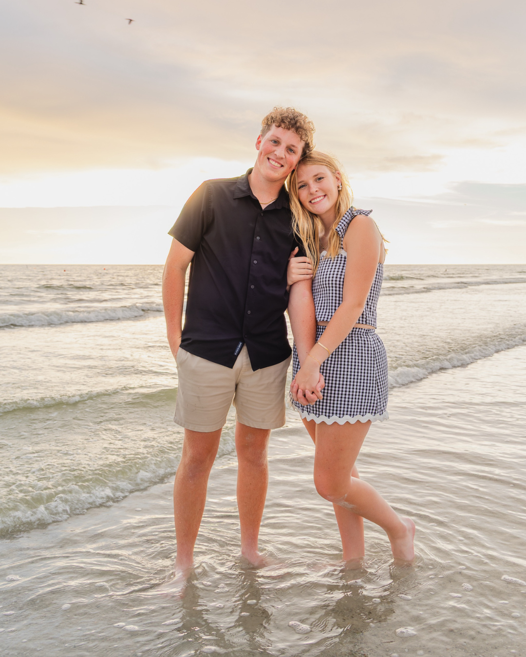 Olivia and Carson standing together in the ocean waves at sunset, smiling and holding hands. Lifestyle couples session portrait.
