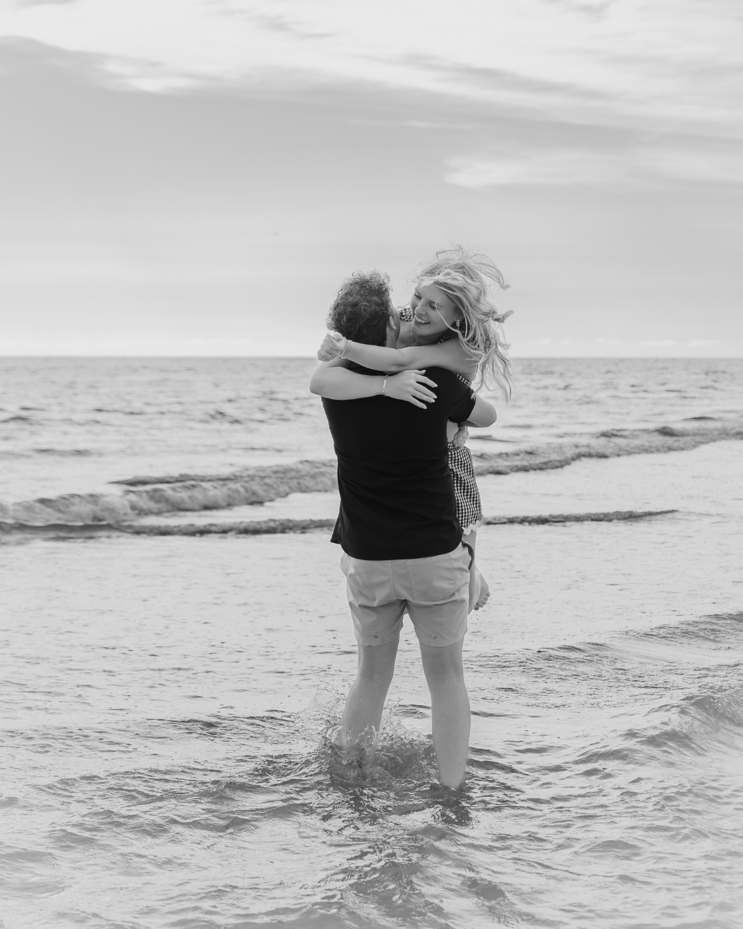 Black and white photo of Olivia and Carson in a full embrace, standing in the ocean surf. Their hair is windblown, showing a passionate moment from their couples session.