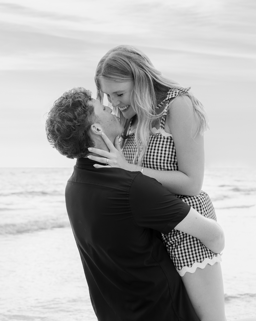 Black and white close-up of Carson lifting Olivia up in the air at the beach, hands on her face. Both are laughing. Intimate moment from a lifestyle couples session.