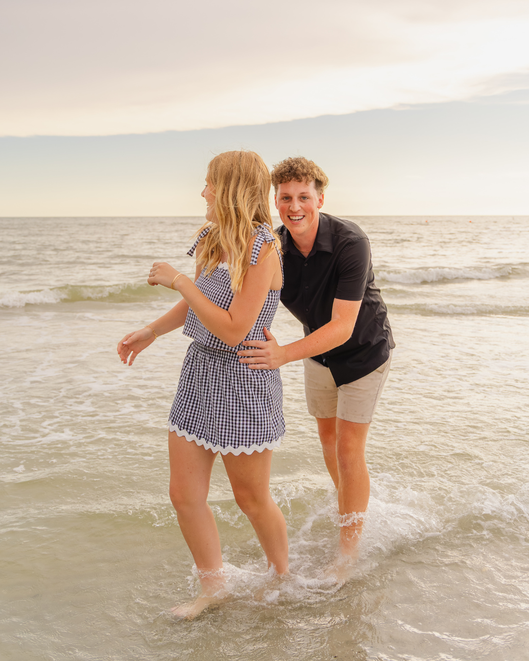 Carson smiling at the camera while walking in the surf with Olivia, who is looking away. A cheerful, candid moment from the beach couples session.