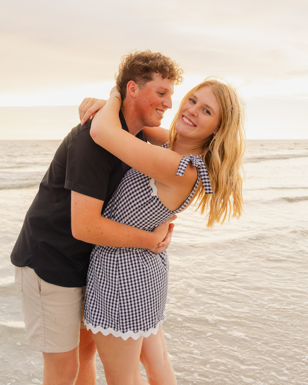 Carson hugging Olivia from behind on the beach during golden hour. Olivia smiles brightly directly at the camera. Romantic couples session photo.