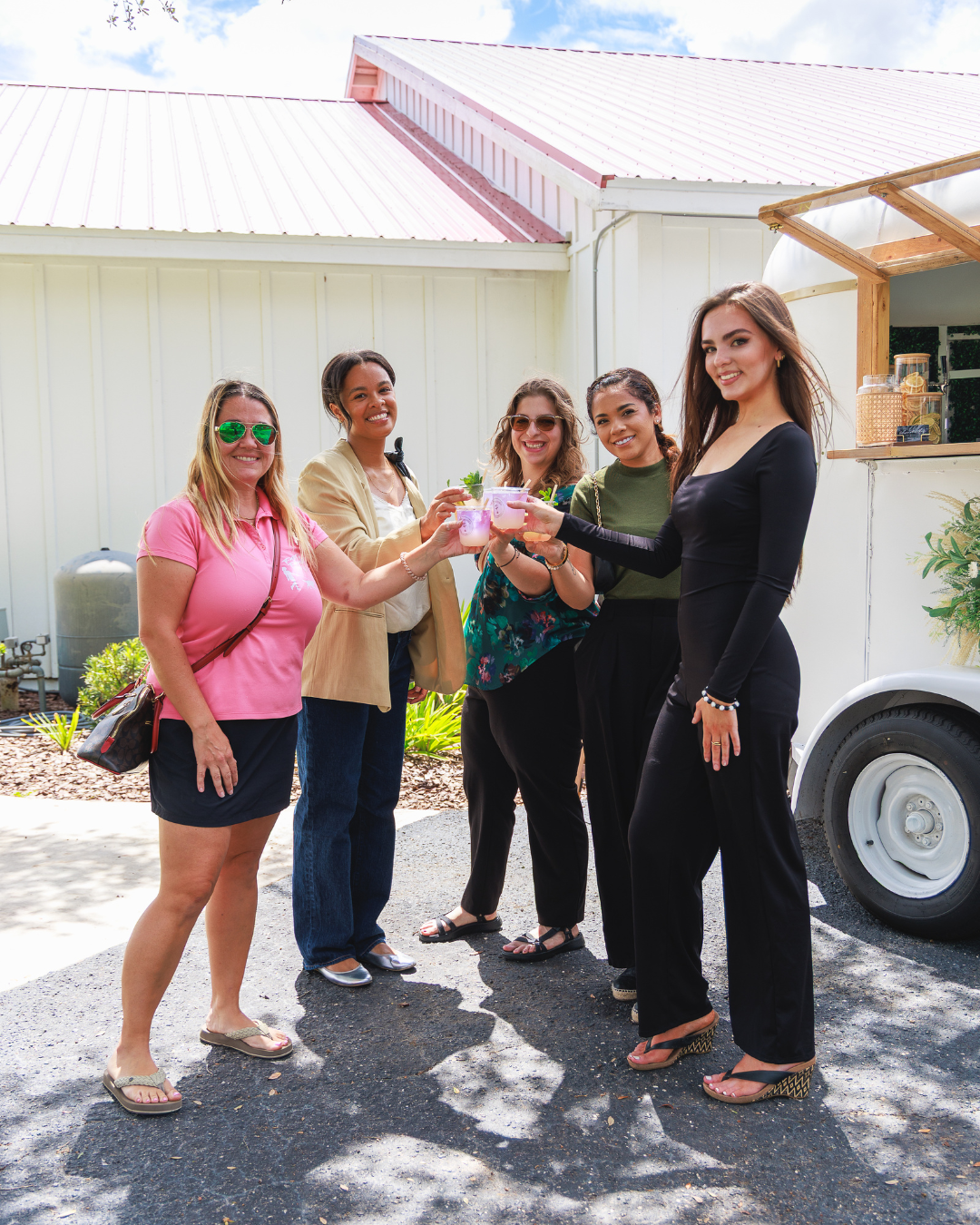 A group of five diverse women smiling and toasting their drinks outdoors in front of a white building and a mobile bar at the United Women’s Network of Florida inaugural women’s networking event. Represents camaraderie and celebration.