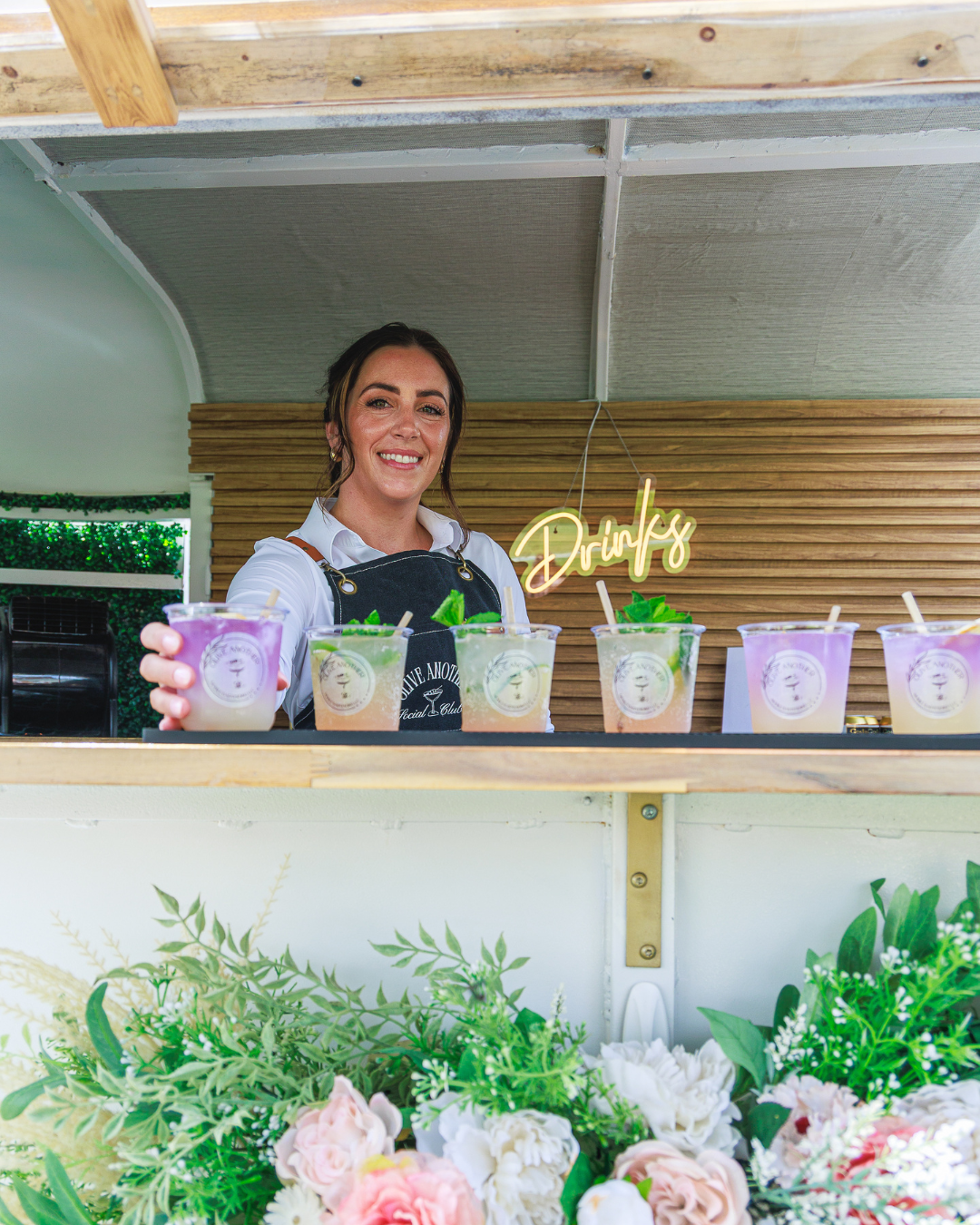 A friendly bartender smiling and serving colorful custom drinks from a stylish mobile bar with a "Drinks" neon sign at the United Women’s Network of Florida inaugural event. Highlights hospitality and refreshments.