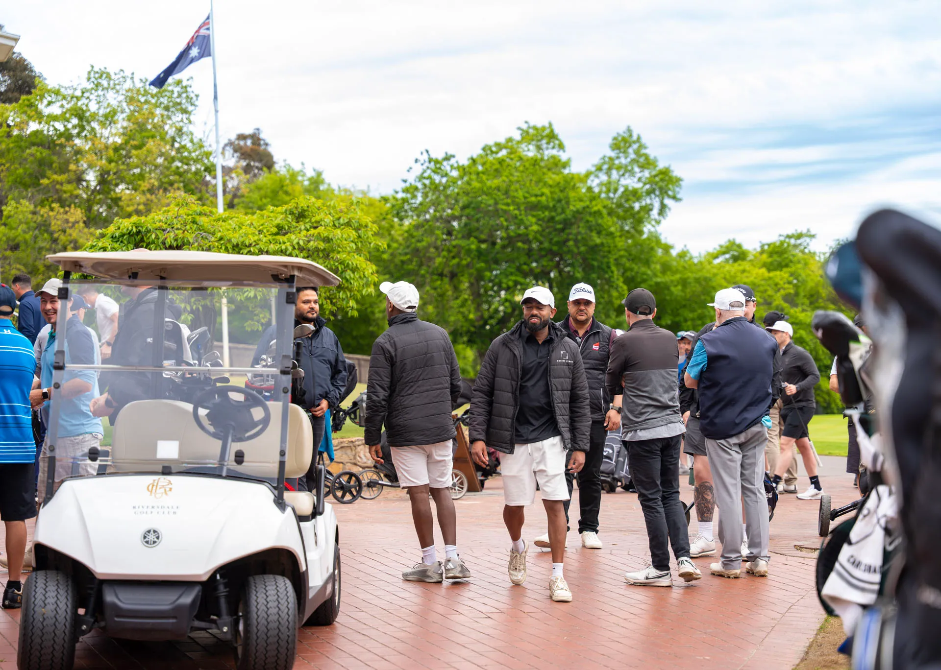 Group of men in casual golf attire gathered around a golf cart and on a brick path at a golf course with trees and an Australian flag in the background.
