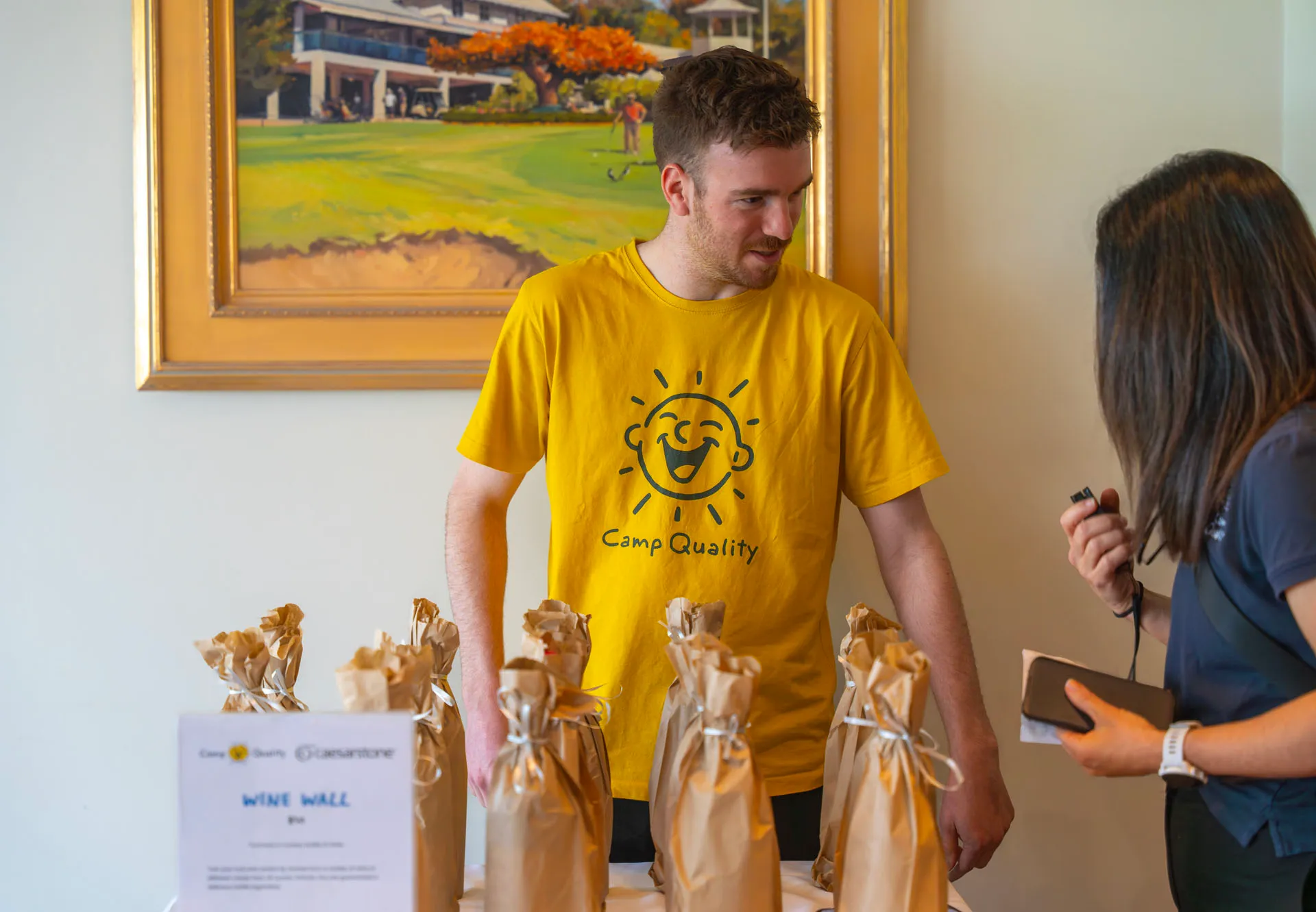 Man in yellow Camp Quality shirt standing behind a table with brown paper-wrapped bottles, talking to a woman holding a phone and microphone.
