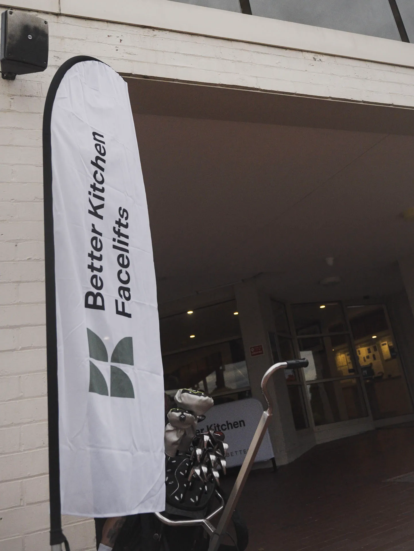 White vertical banner reading 'Better Kitchen Facelifts' next to a golf bag with clubs outside a building entrance.