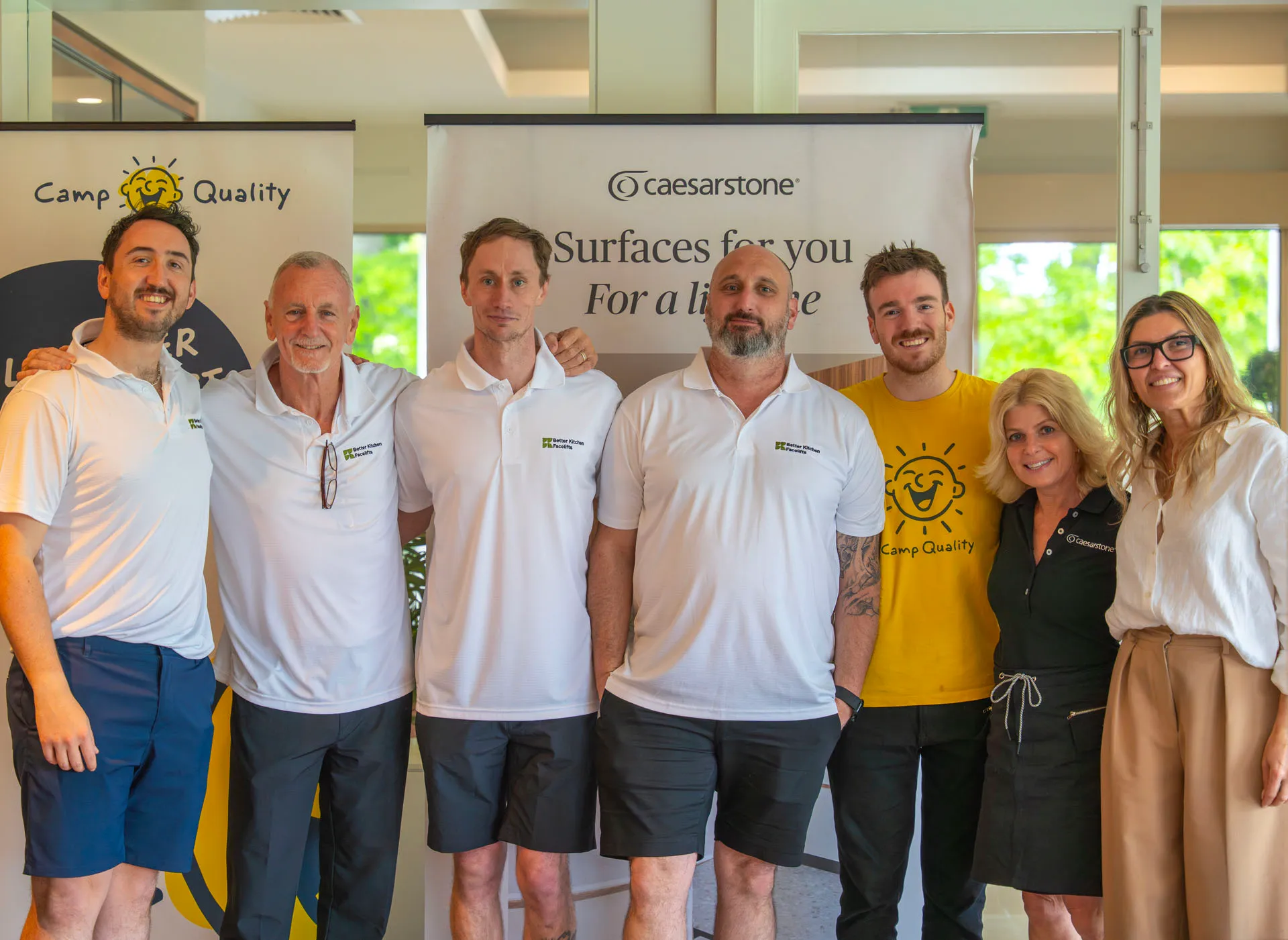 Group of seven people standing indoors in front of promotional banners, smiling and posing together; four men in white polo shirts and black shorts, one man in a yellow Camp Quality shirt, and two women, one in a black dress and one in beige pants and white blouse.