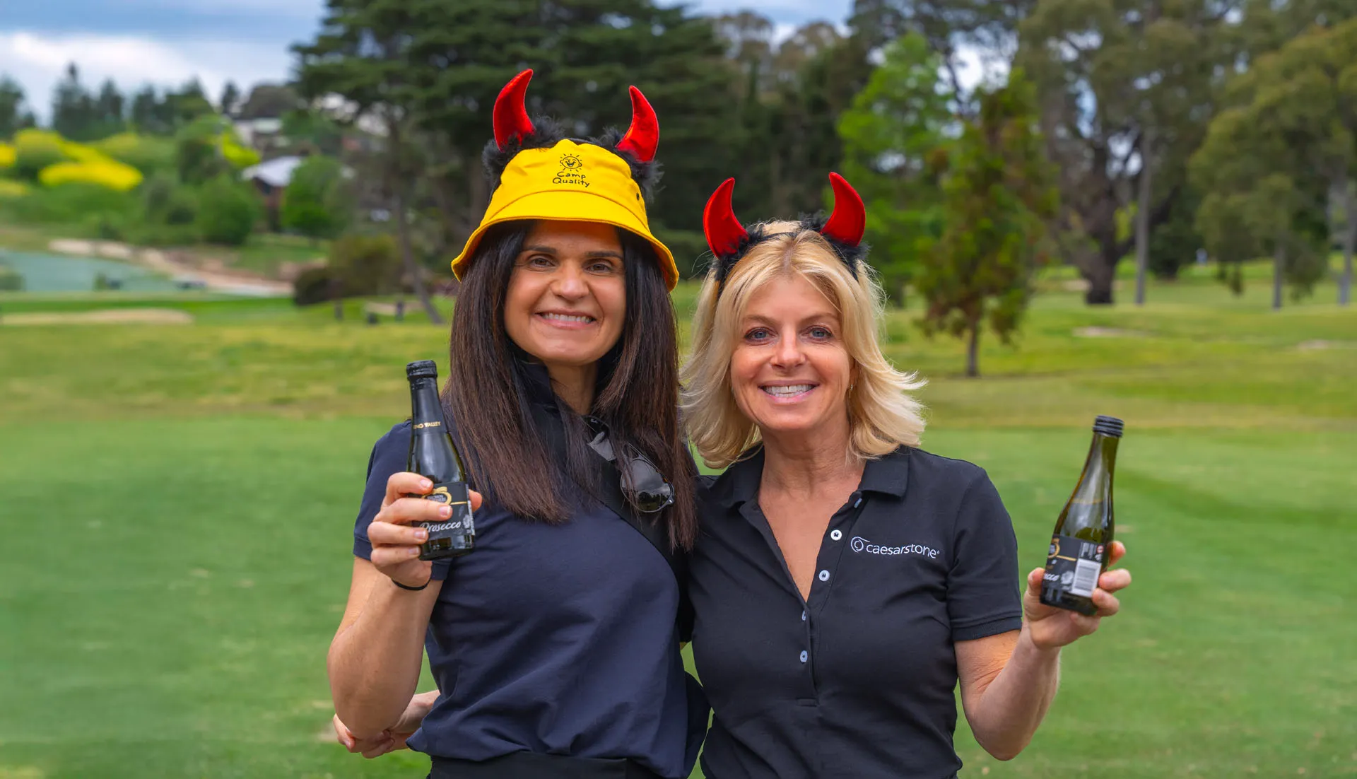 Two smiling women outdoors wearing red devil horn accessories and holding small bottles of Prosecco.