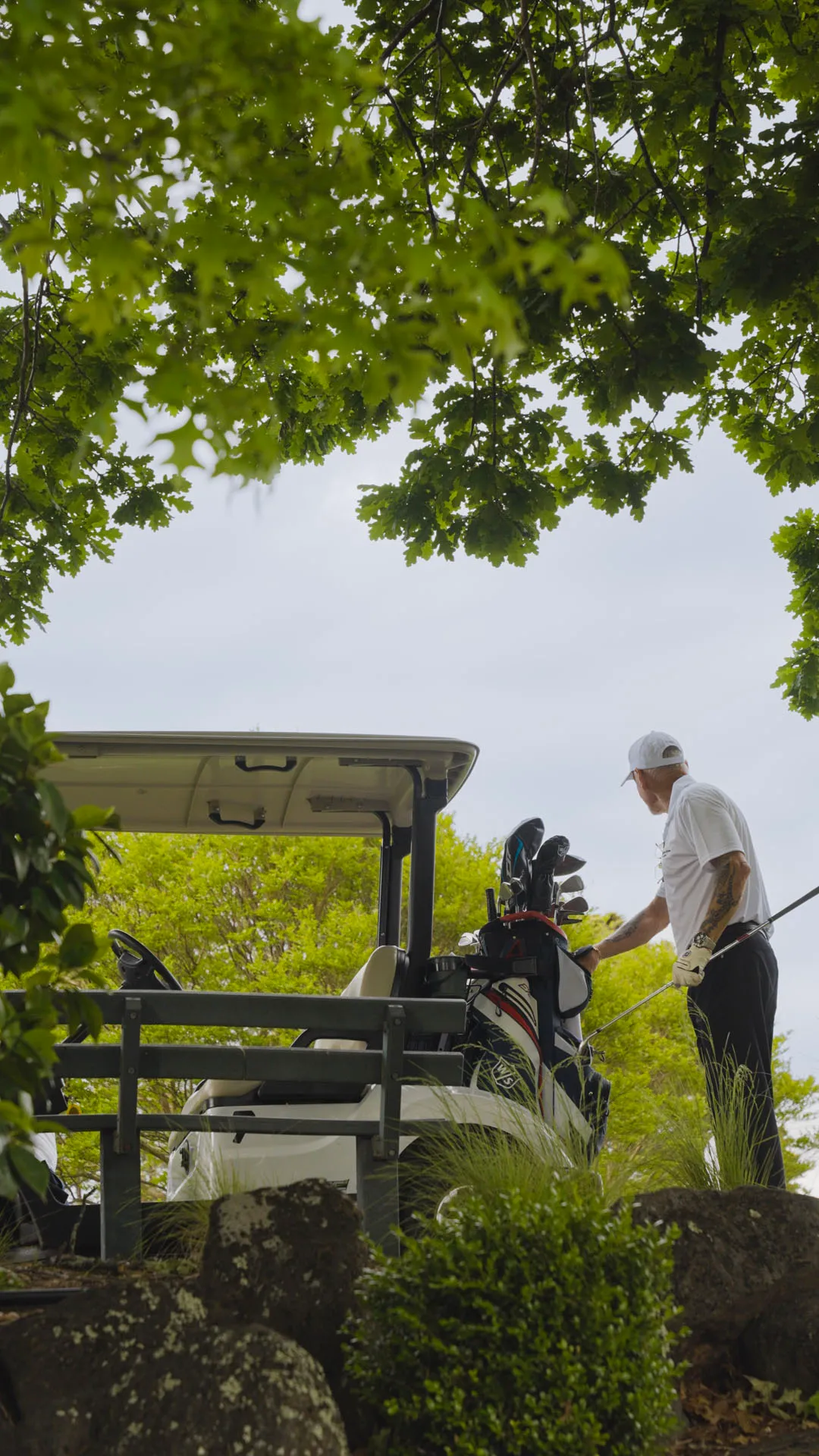 Man in white cap and gloves standing next to a golf cart with golf clubs, surrounded by green trees and bushes.