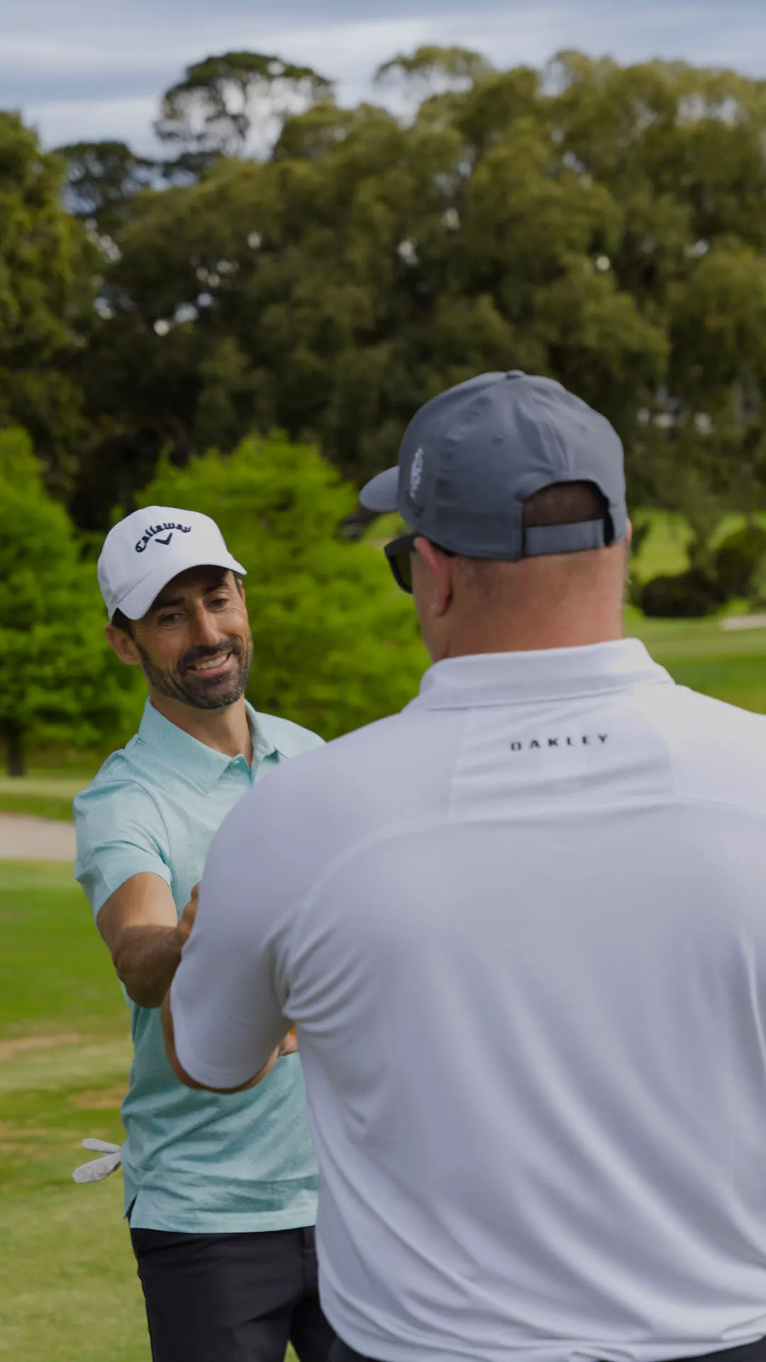 Two men on a golf course shaking hands, one wearing a light blue shirt and white Callaway cap, the other wearing a white Oakley shirt and gray cap.