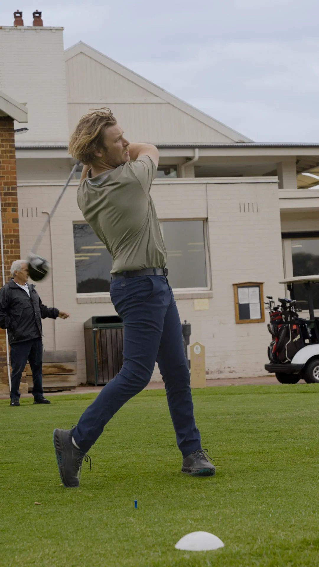 Man in green shirt and navy pants swinging a golf club on a golf course with a building and another man in the background.