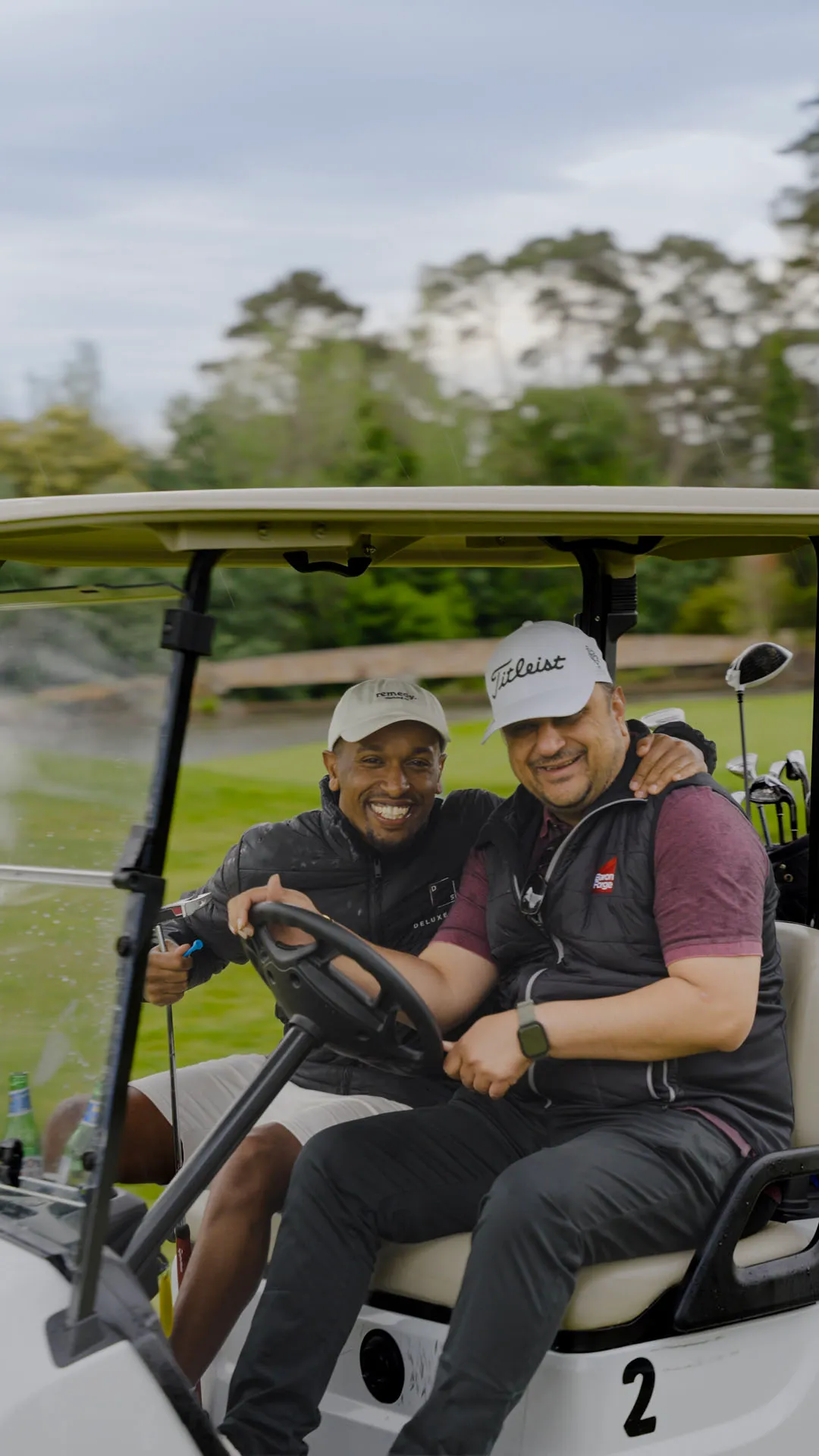 Two men smiling and sitting close together in a golf cart on a golf course, one holding a golf club.