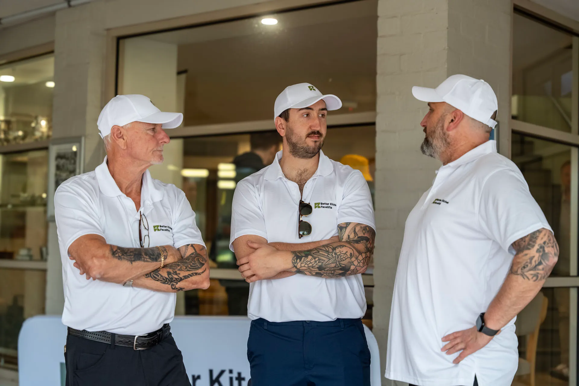 Three men wearing white polo shirts and white caps with company logos, standing with arms crossed and engaged in conversation indoors.