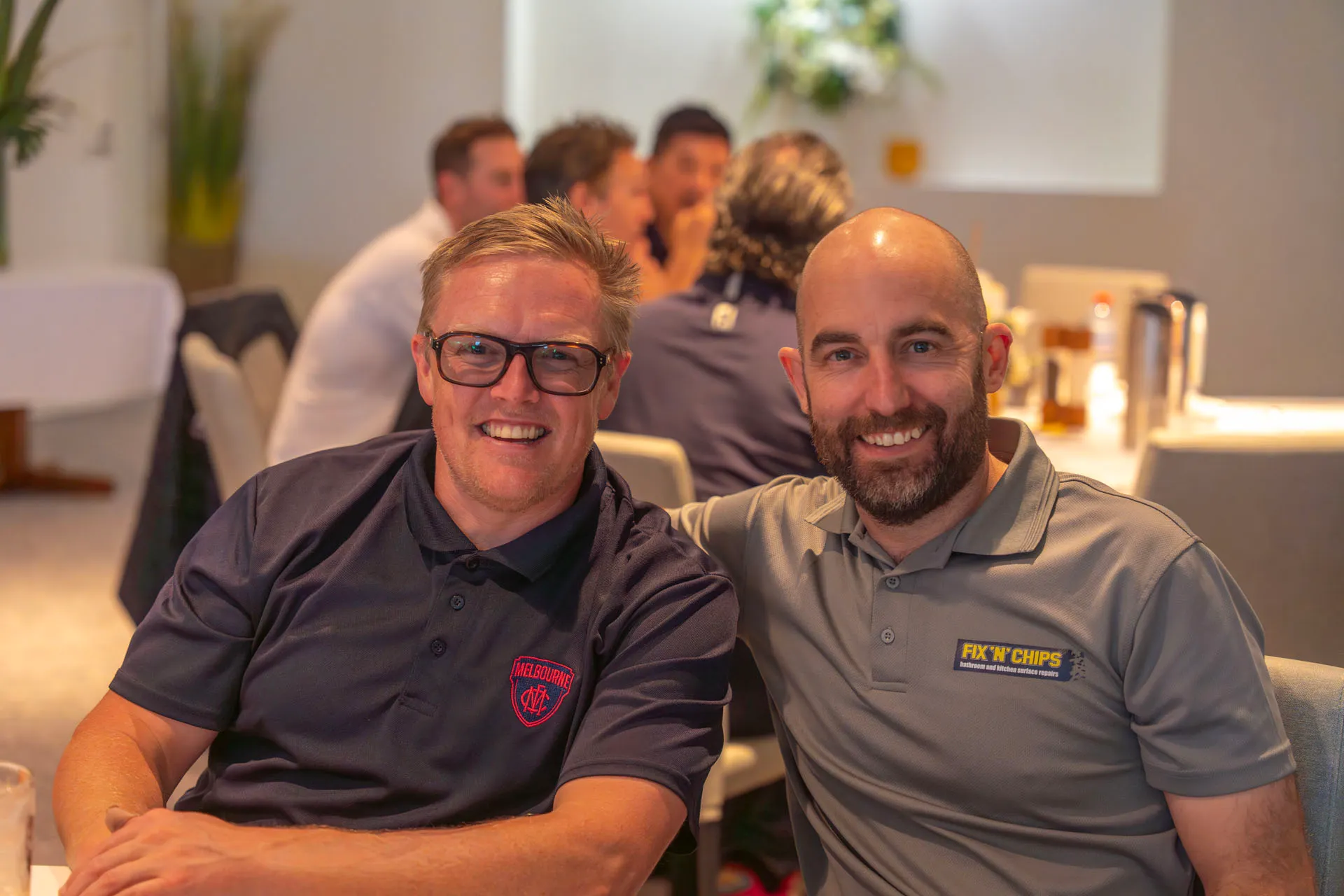 Two men smiling and sitting closely together in a well-lit room with people and tables in the background.