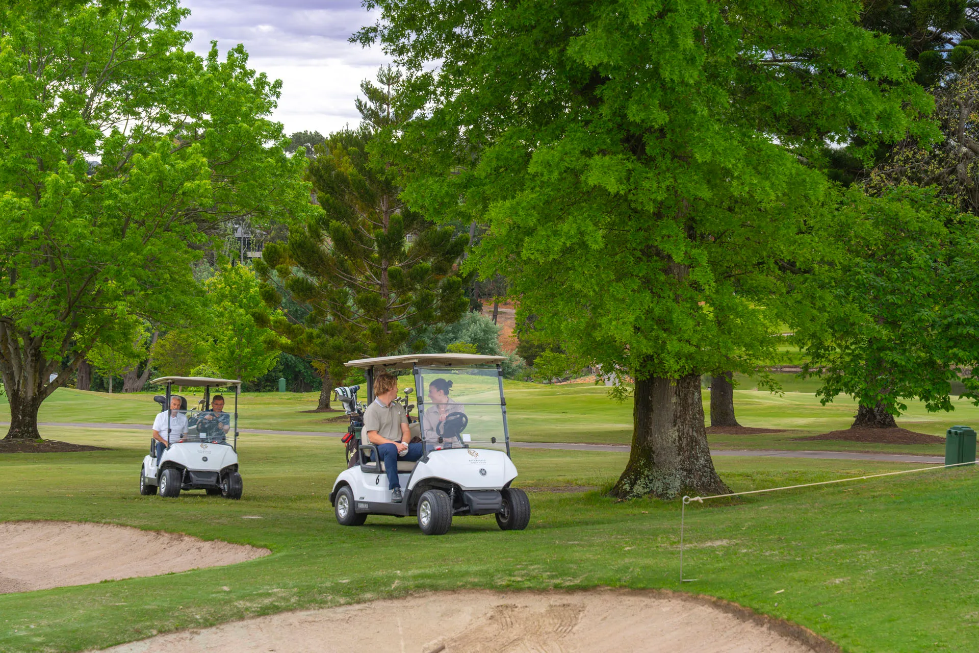 Two golf carts on a green golf course with four people, surrounded by large trees and sand bunkers.