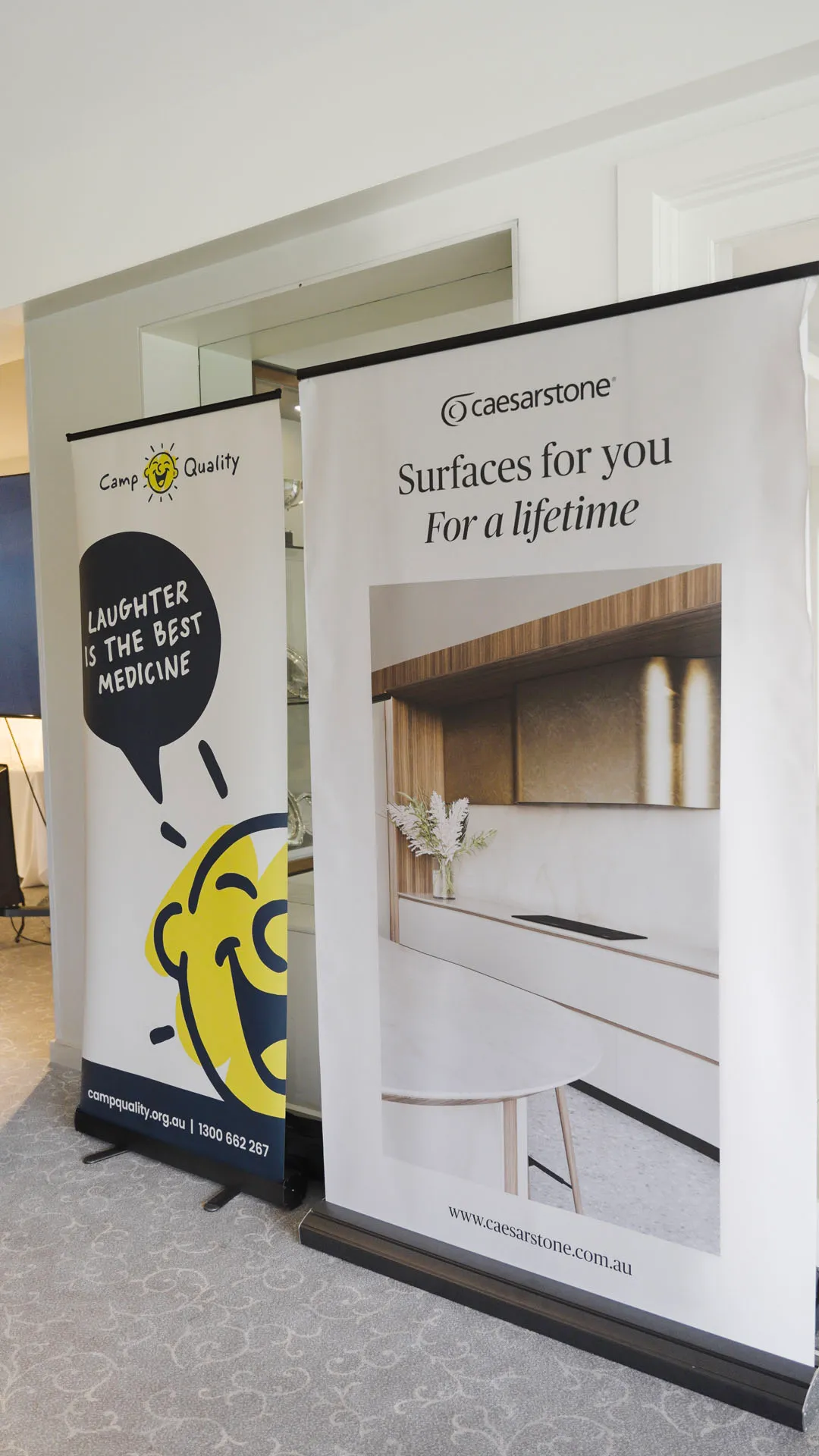 Two standing banners side by side: one with a smiling yellow face and text 'Laughter is the best medicine,' the other showing a modern kitchen with Caesarstone surfaces and the slogan 'Surfaces for you For a lifetime.'