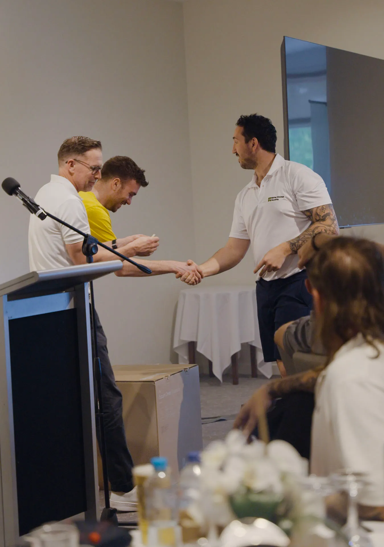 Man in white shirt shaking hands with man in yellow shirt at an indoor event with a microphone and podium nearby.