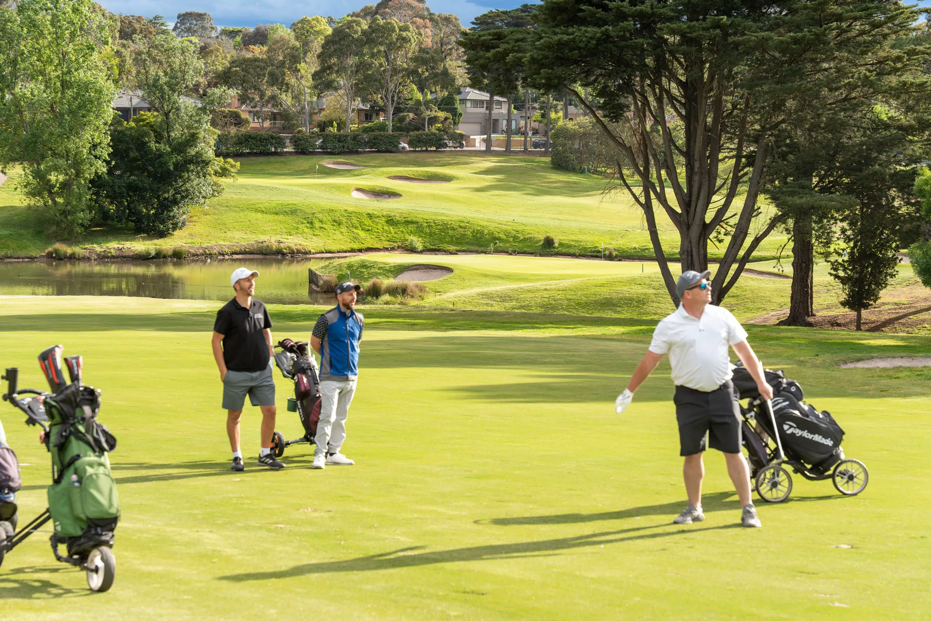 Three men on a golf course near bags and trolleys, with one man swinging a golf club.