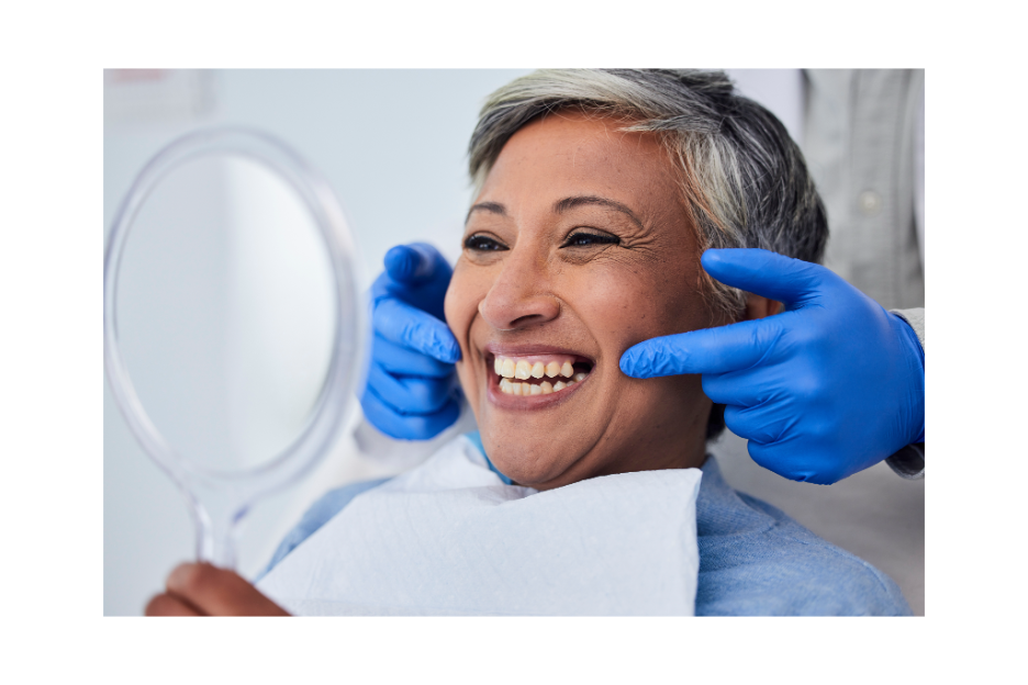 Smiling dental patient holding a hand mirror while a clinician in blue gloves points to her gum line during a cosmetic smile consultation.