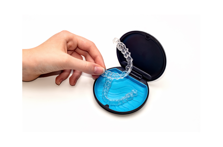 Hand lifting a clear dental aligner from an open round storage case, illustrating removable Invisalign-style trays used to straighten teeth.
